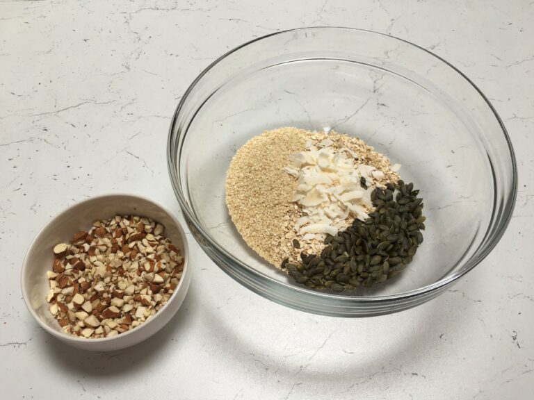 Preparazione granola fatta in casa: ciotola grande di vetro con fiocchi di avena, cocco e semi e ciotolina con mandorle a pezzetti